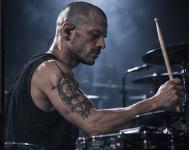 Pulse playing drums on stage, tattoos visible on his arm.